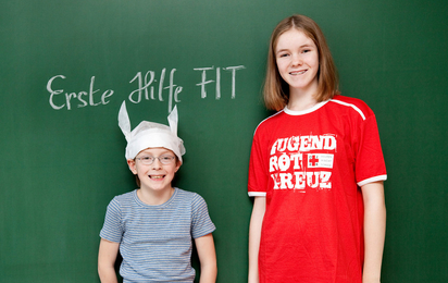 Zwei Kinder stehen vor einer Tafel, auf der "Erste Hilfe FIT"