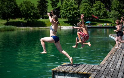 Kinder springen mit Anlauf von einem Steg in einen Badesee.